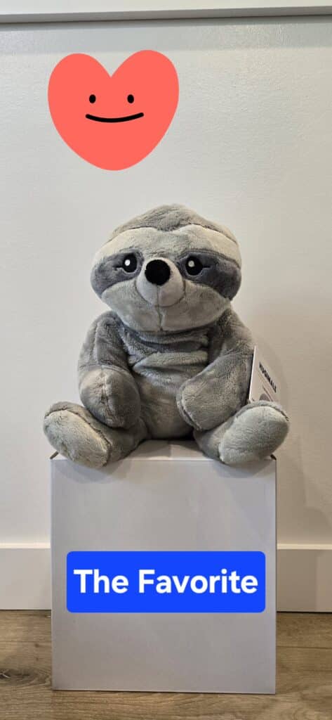The Hugimal Sam the Sloth weighted sensory plush toy sitting on a box, showing its soft gray fur and friendly expression.